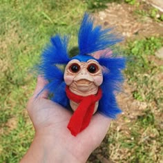 a hand holding a small troll doll with blue hair and red scarf on it's head