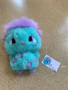 a blue stuffed animal with purple hair sitting on the floor next to a sticker