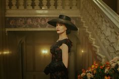 a woman in a black dress and hat is standing on the stairs with flowers behind her