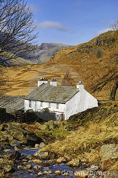 a small white house sitting on the side of a hill next to a stream and trees