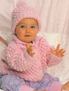 a baby wearing a pink knitted sweater and matching hat is sitting on a bed