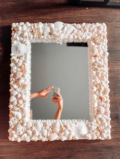 a hand holding a cell phone in front of a mirror with shells on the frame