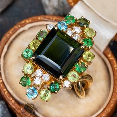 This impressive cocktail ring is centered with a cut cornered, rectangular step cut tourmaline, weighing 7.23 carats, in a four-prong setting. The top of the ring is prong set with eight (8) round brilliant cut diamonds, six (6) round mixed cut tsavorites and peridots and two (2) round mixed cut blue topaz and tourmaline. The ring measures 27.0mm at the top, rises 9.3mm above the finger, tapering to 2.5mm wide and 0.7mm thick at the base of the shank. This ring is currently a size 6.75. Tourmaline Rings, 2nd Wedding, Colored Stone Rings, Gold Cocktail Ring, Tourmaline Ring, Unique Gemstones, Green Tourmaline, Rings Jewelry, Brilliant Diamond