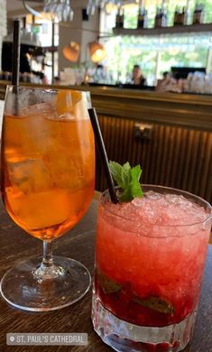 two cocktails sitting on top of a wooden table next to each other in front of a bar