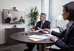 two business people sitting at a table in an office setting with a television on the wall