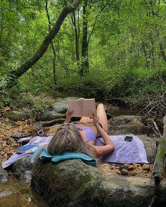 a woman laying on top of a rock next to a river while reading a book