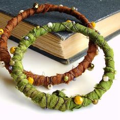 two bracelets made out of green and brown fabric with beads on them next to a book