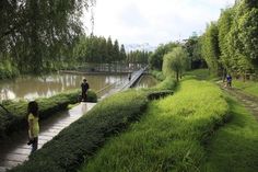 two people are walking along a path near the water and some bushes on either side