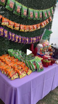 a purple table topped with lots of bags of food next to a green wall covered in plants