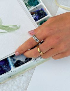 a woman's hand with two rings on her finger next to an open box of watercolors