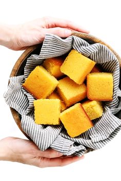 two hands holding a wooden bowl filled with squares of yellow cake on top of a gray and white striped napkin