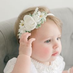 A baby girl with blue eyes and light hair wearing the Aria Christening Gown and the Aria Flower Headband, gently touching the headband while sitting on a grey chair. Baggy Sweaters, Flowers And Greenery, Heirloom Gifts, Baby Comforter, Create Outfits, Flower Headband, Soft Hands, Soft Hand, Flowers And Leaves