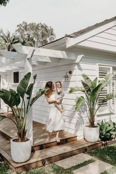 a woman holding a baby standing on a porch next to a house with potted plants