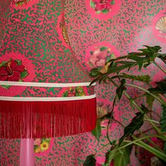 a pink and green wallpaper with red fringe lampshade next to potted plant