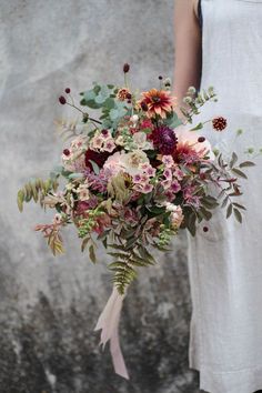 a woman holding a bouquet of flowers in front of a stone wall with leaves and foliage