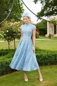 a woman in a blue dress and hat standing on the grass with bushes behind her