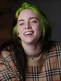 a woman with green hair is smiling and wearing a plaid sweater in front of a dark background