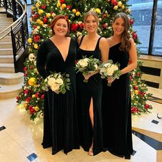 three women standing in front of a christmas tree wearing long dresses and holding bouquets