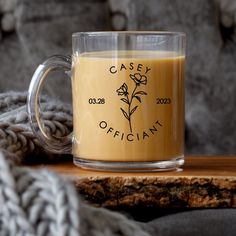 a glass mug with a flower on it sitting on a wooden table next to a gray sweater