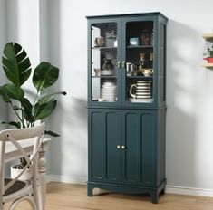 a tall green cabinet with glass doors in a room next to a chair and potted plant