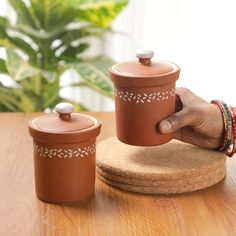 a person holding a brown ceramic container on top of a wooden table next to a potted plant