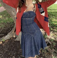 a woman standing in front of a tree wearing a red cardigan and blue denim dress