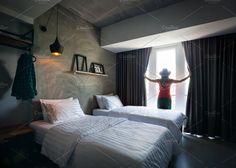 a woman standing at the window in a hotel room with two beds and one is holding her arms up