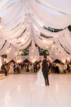 a bride and groom dance under white draping at their wedding reception with chandeliers hanging from the ceiling