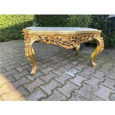 an ornate gold coffee table with glass top on cobblestone pavement in front of shrubbery