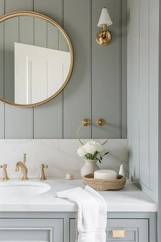 a bathroom with gray walls and white countertops, gold fixtures and a round mirror