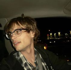 a man wearing glasses sitting in the back seat of a car with city lights behind him