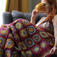 a woman sitting on a couch drinking out of a cup while holding a crocheted blanket