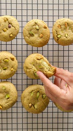 a hand holding a cookie in front of several cookies on a cooling rack with nuts and pistachios