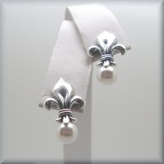 "Sterling silver fleur de lis stud earrings are accented with a glowing white Swarovski elements half-drilled pearl. Classic, beautiful and unusual. This symbol has traditionally been used to represent French royalty, and in that sense it is said to signify perfection, light, and life. Fleur de lis translates to \"flower of the lily\" in English. The earrings are about 7/8\" long (1.8 cm) from the top of the fleur de lis to the bottom of the pearl. Setting, earnuts and earring components are all Formal White Sterling Silver Flower Earrings, Classic White Flower Earrings For Pierced Ears, White Classic Flower Earrings For Pierced Ears, Classic Flower Earrings, Classic White Flower Earrings, White Classic Flower Earrings, Classic White Flower Earrings For Anniversary, Classic White Flower Earrings For Wedding, White Classic Flower Earrings For Anniversary