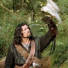 a man holding an owl on his arm in the woods with trees and plants behind him