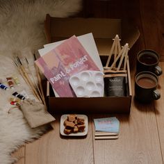 a box filled with lots of crafting supplies on top of a wooden floor next to two mugs