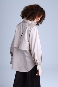 The beige caped shirt Diva is a unique creation that blends classic elegance with a modern twist. This button-down shirt features a cape-like design that gracefully drapes over the shoulders and back, creating a stunning winged effect. The puffy long sleeves with bell-shaped cuffs add a touch of refinement to this exquisite piece. Crafted from premium cotton satin, the Diva Beige shirt is not only luxurious but also comfortable to wear all day long. Its adaptable design allows you to experiment with different bottom options, enabling you to create a one-of-a-kind ensemble that reflects your personal style. Elegant Cape Top For Spring, Elegant Spring Cape Top, Elegant Oversized Beige Tops, Chic Neutral Fall Shirt, Elegant Oversized Fall Tops, Elegant Collared Blouse In Neutral Color, Elegant Neutral Shirt For Spring, Elegant Batwing Sleeve Top For Workwear, Elegant Batwing Sleeve Top For Work