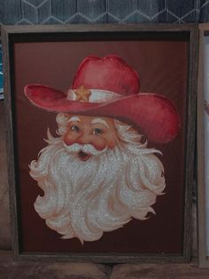 a painting of a santa claus wearing a red cowboy hat and white beard in a wooden frame