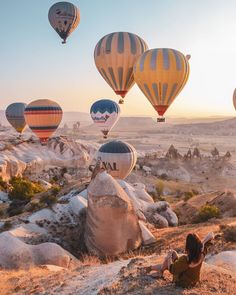 several hot air balloons in the sky above some rocks and trees, with a woman sitting on the ground looking at them