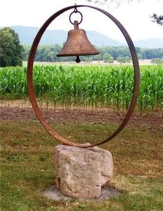 a metal bell sitting on top of a rock in the middle of a field next to corn