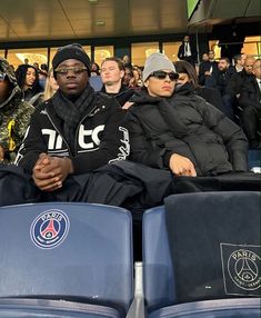 two men sitting in the stands at a sporting event, one is wearing sunglasses and the other wears a beanie