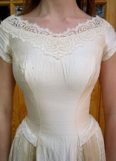 a woman wearing a white dress with lace on the neck and sleeves, standing in front of a wooden door