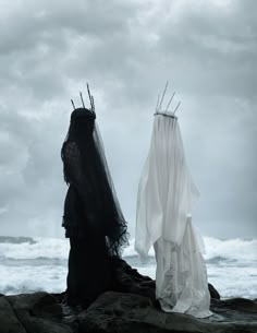 two people dressed in black and white standing on rocks near the ocean under cloudy skies