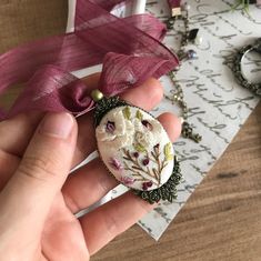 a hand holding a brooch with flowers on it and a purple ribbon around it