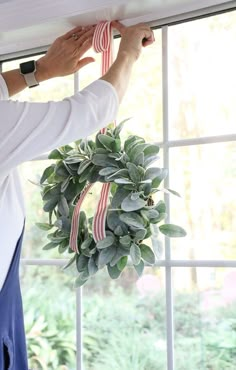 a woman hanging a wreath on the side of a window with red and white ribbon