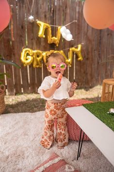 a baby girl wearing sunglasses standing in front of a table with balloons and streamers