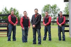 five men standing in front of a black and white barn with red vests on