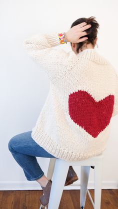 a woman sitting on top of a stool holding her hands to her head while wearing a heart sweater