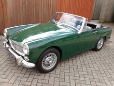 an old green sports car is parked in front of a wooden wall and brick walkway