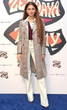 a woman with long hair standing in front of a wall wearing a coat and white pants